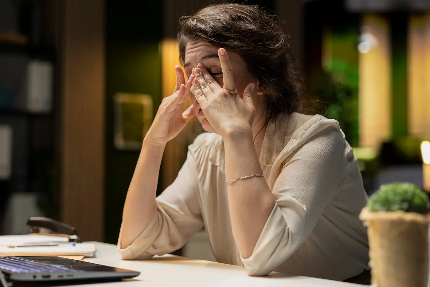 A woman at her desk in the evening, rubbing her eyes with both hands